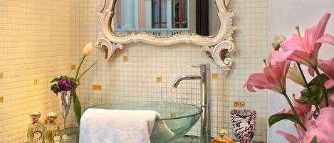 Un baño con lavamanos de vidrio, toallas blancas, un espejo con un marco ornamentado y un florero con flores.
