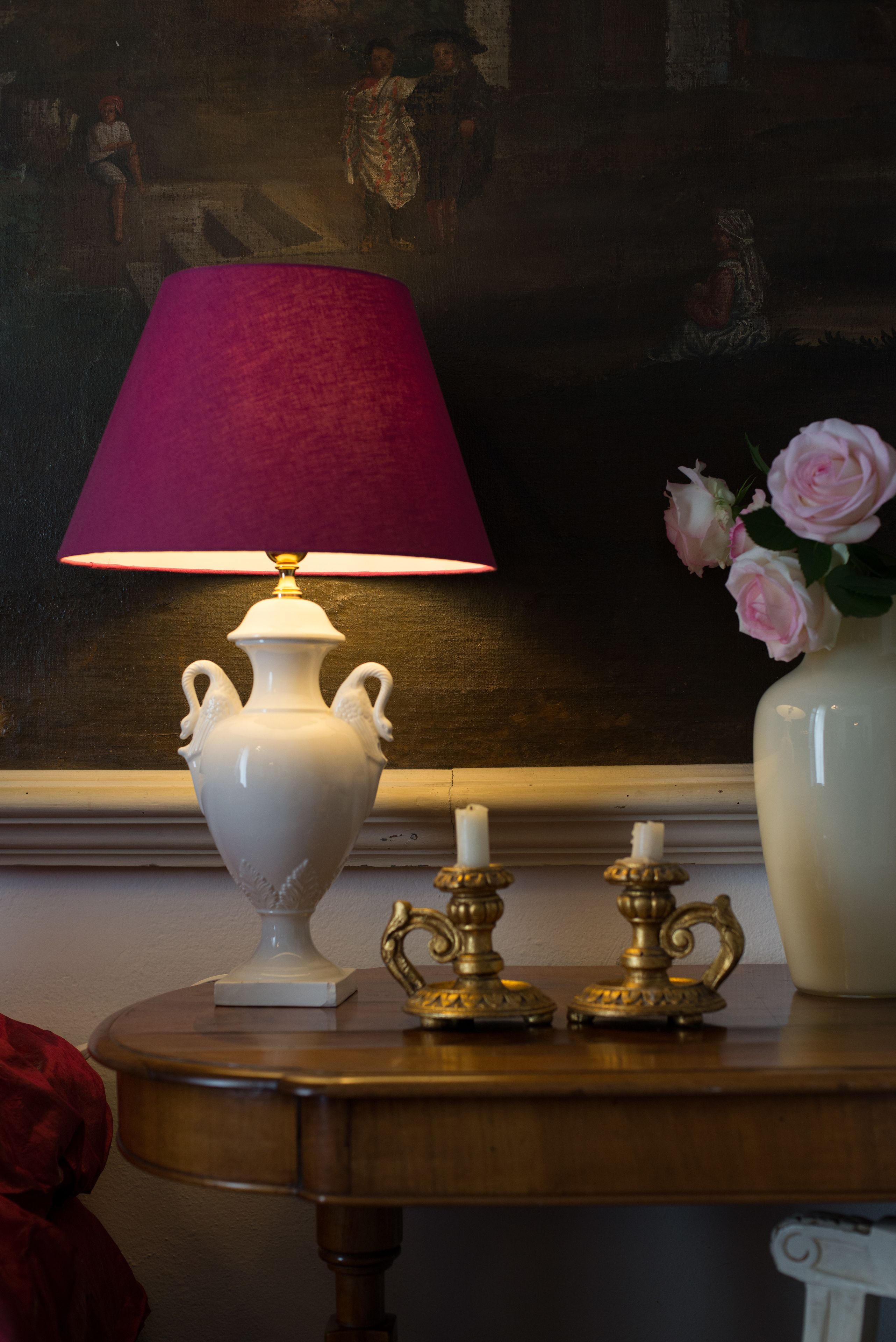 Una mesa con un jarrón blanco con rosas rosadas, una lámpara blanca con pantalla roja y dos candelabros de bronce.