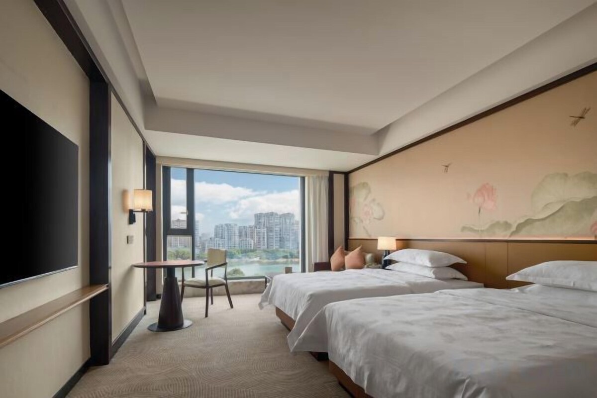 A hotel room with two beds, a desk, a chair, and a large window offering a city view.
