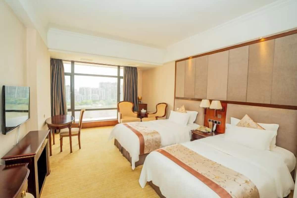 A hotel room with two beds, a desk, a chair, a TV, and a large window with curtains.
