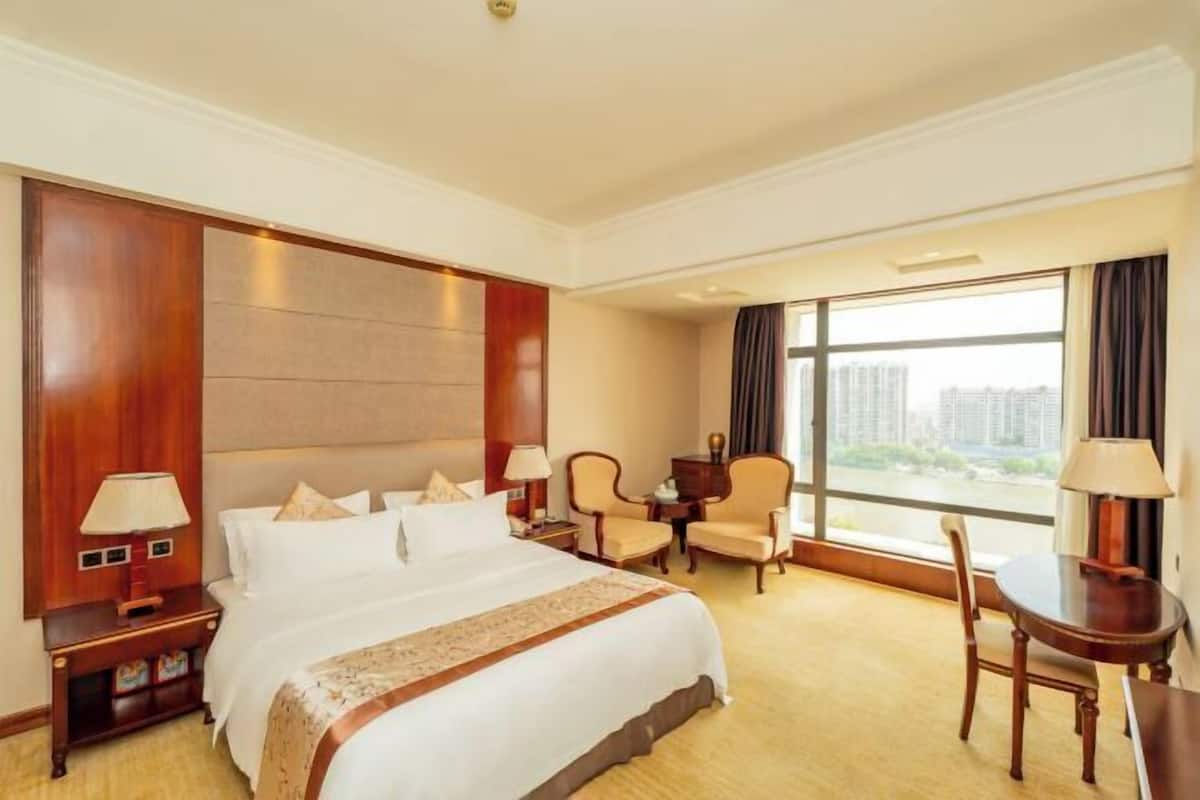 A hotel room with a large bed, a desk, two chairs, a window with a view of buildings, and a nightstand with a lamp.