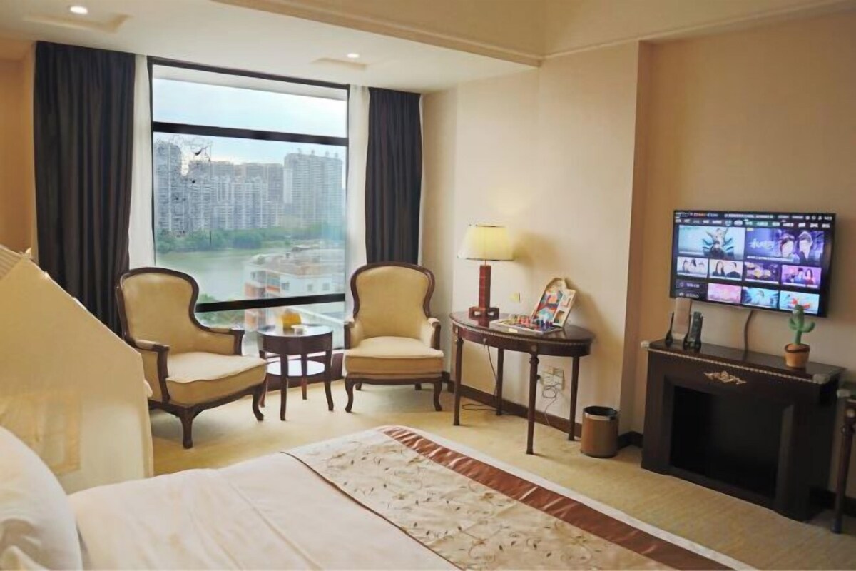 A hotel room with a bed, two armchairs, a small table, a TV, and a view of buildings through the window.
