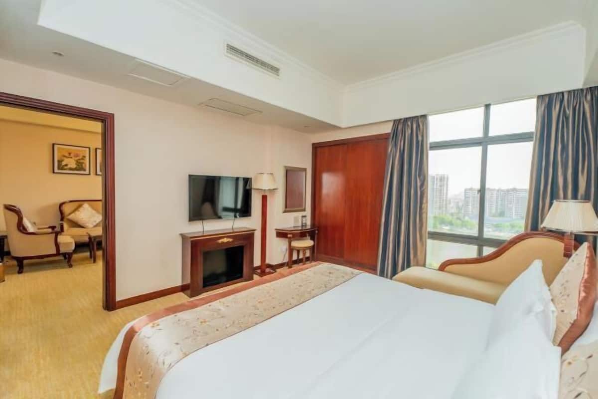 A hotel room with a large bed, a television, a desk with a lamp, and a view of the city through the window.