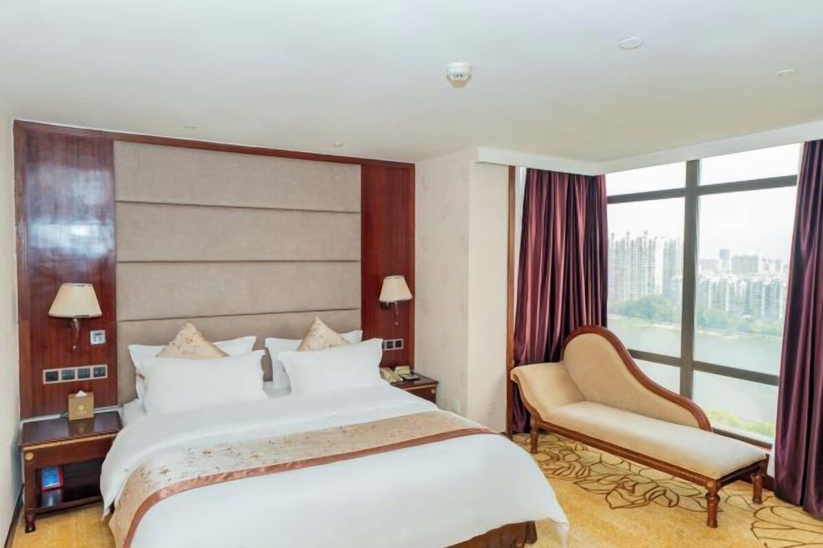 A hotel room with a large bed, a nightstand, a chair, and a view of the city through large windows.