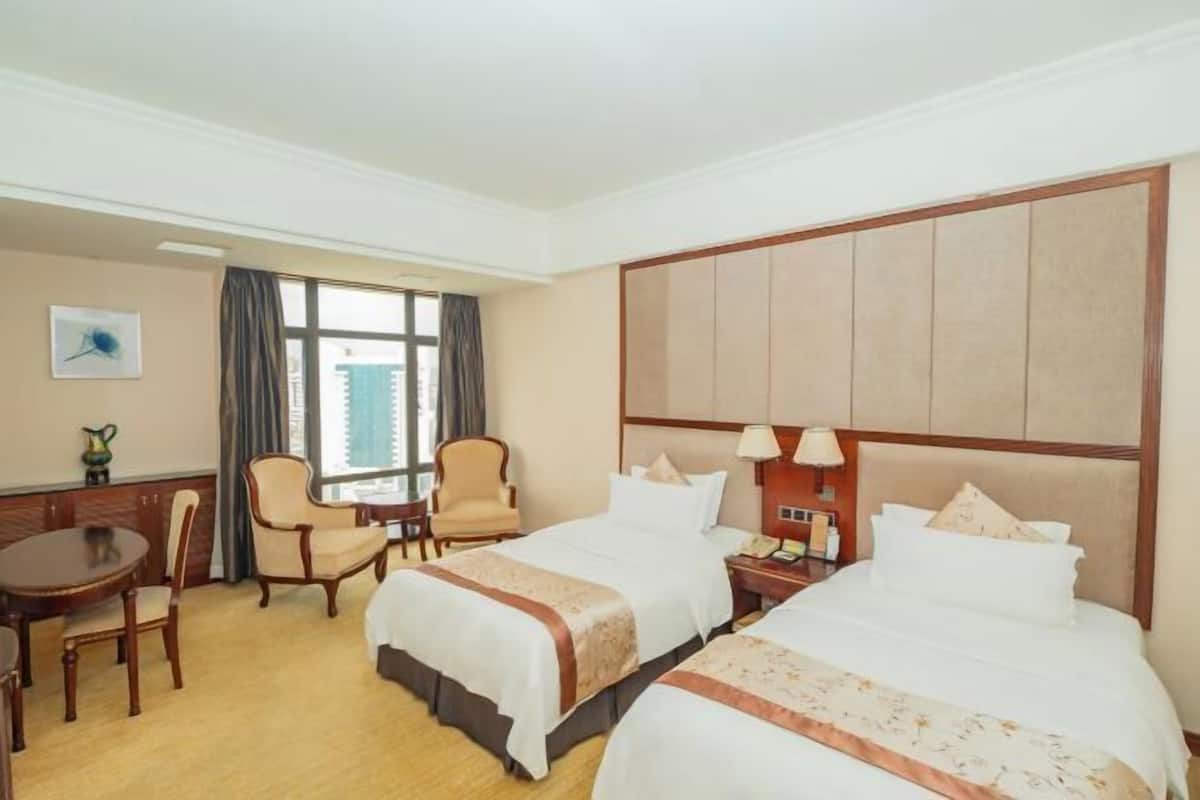 A hotel room with two beds, a desk with a chair, a window with curtains, and a wall-mounted lamp.