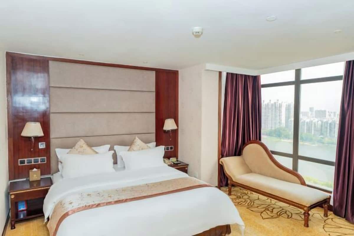 A hotel room with a large bed, a nightstand, a chair, and a view of the city through large windows.