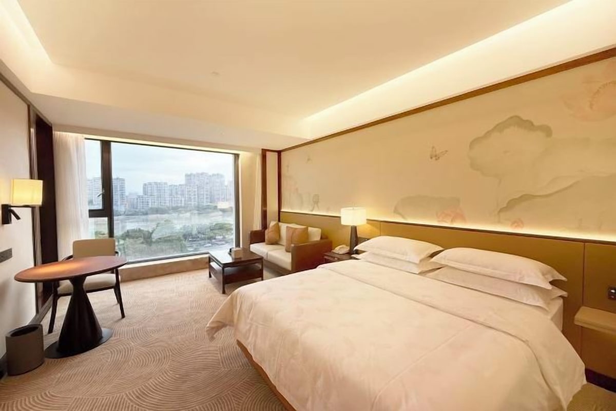 A hotel room with a large bed, a desk, a chair, and a view of the cityscape through the window.
