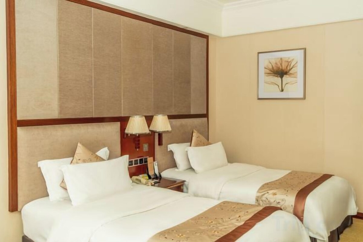 A hotel room with two beds, a large headboard, a nightstand with lamps, and a framed picture on the wall.