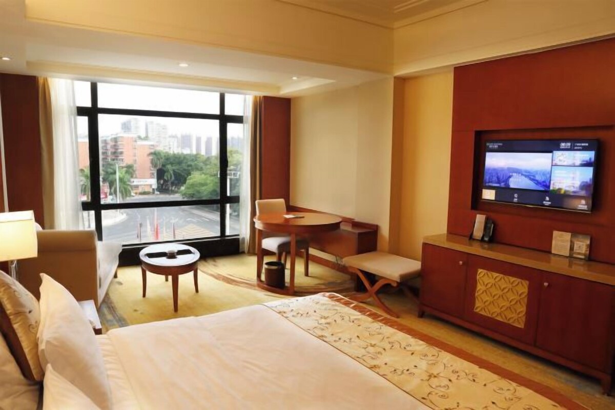 A hotel room with a bed, a desk, a chair, a TV, and a view of the city through large windows.