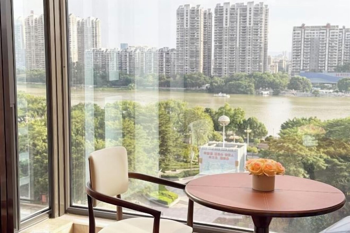 A room with a view of a river and high-rise buildings, a round table with a vase of flowers, and a chair.
