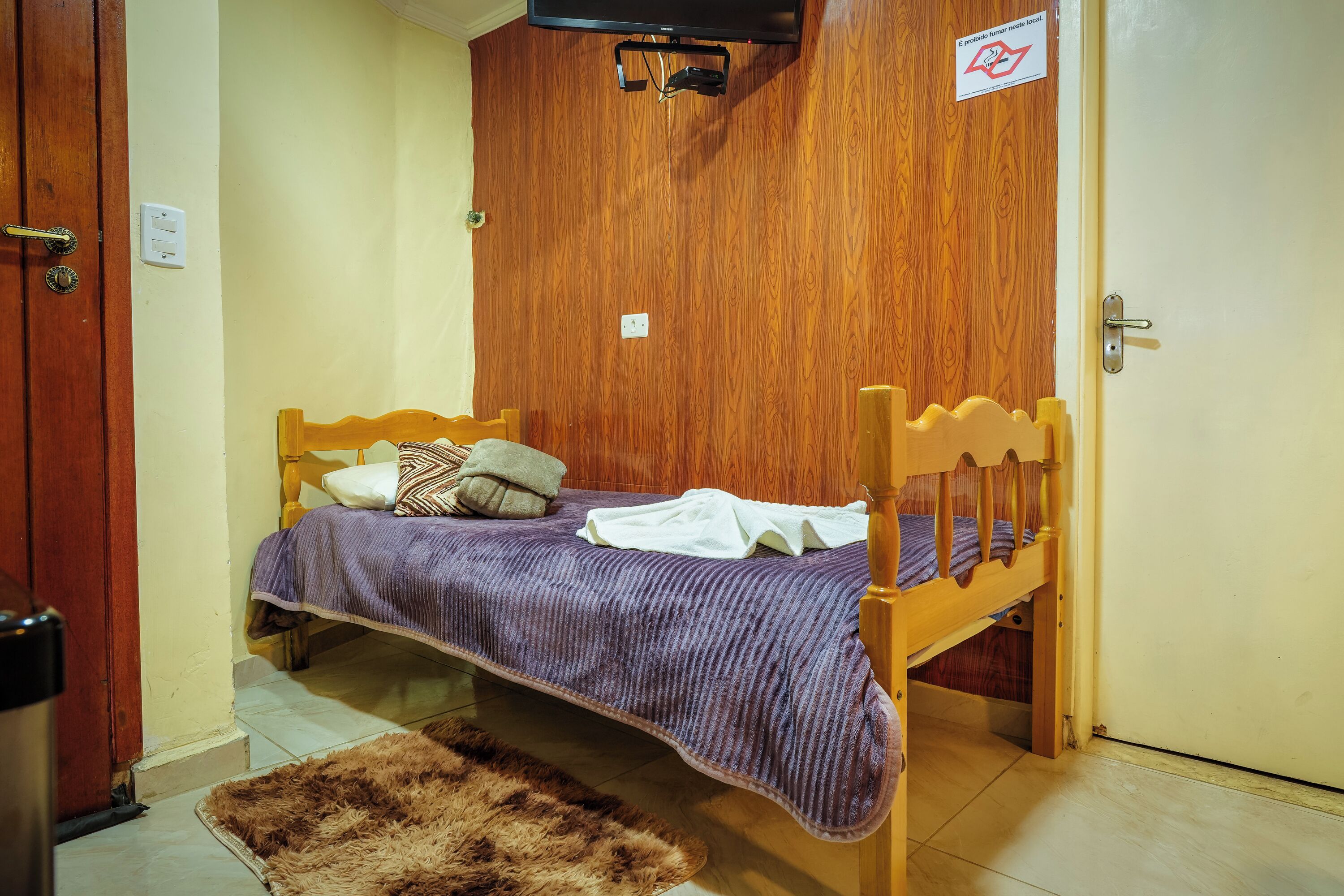 Une petite chambre avec un lit simple, une tête de lit en bois, une couvre-lit violette et un tapis marron sur le sol.