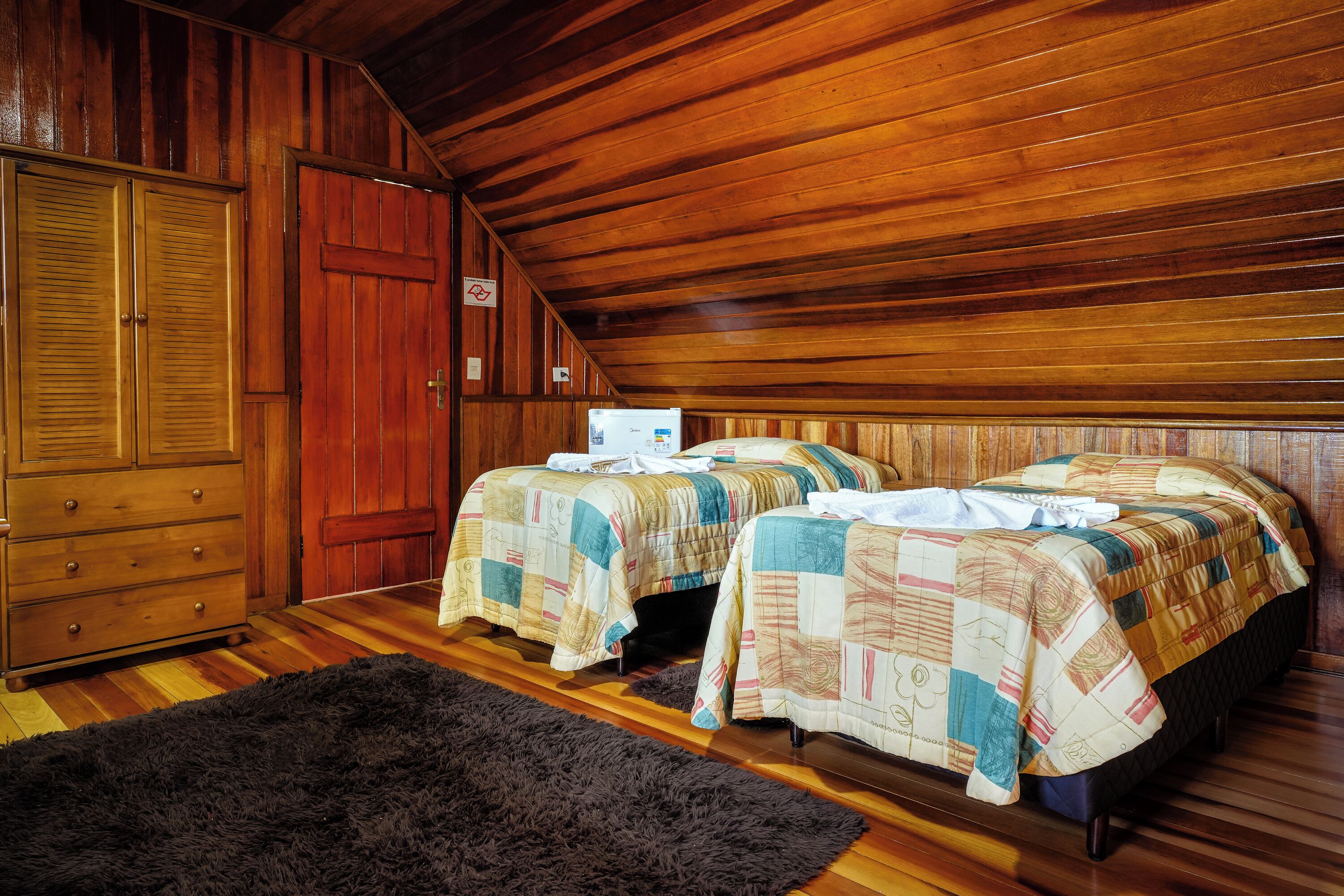 A room with two beds, wooden walls, a large wardrobe, and a black rug.