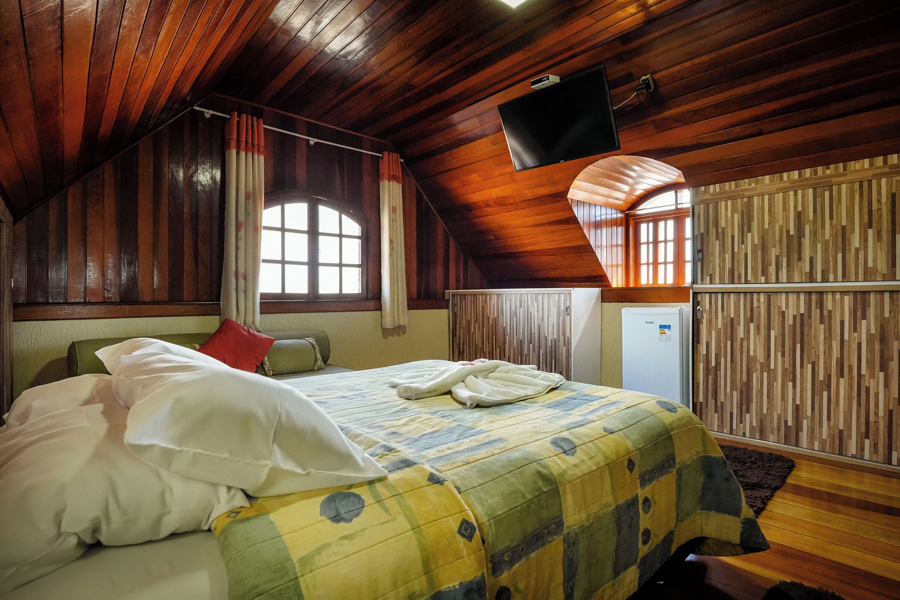 Una habitación acogedora con techo de madera, cama con colcha colorida, televisor de pantalla plana en la pared y ventana con cortinas.