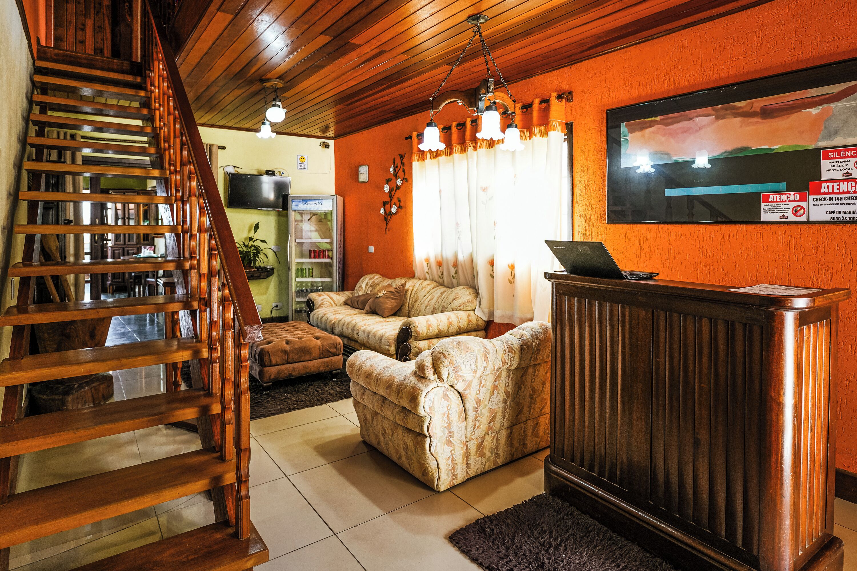 Um saguão de hotel com escada de madeira, balcão de recepção, sofá e tela de televisão.