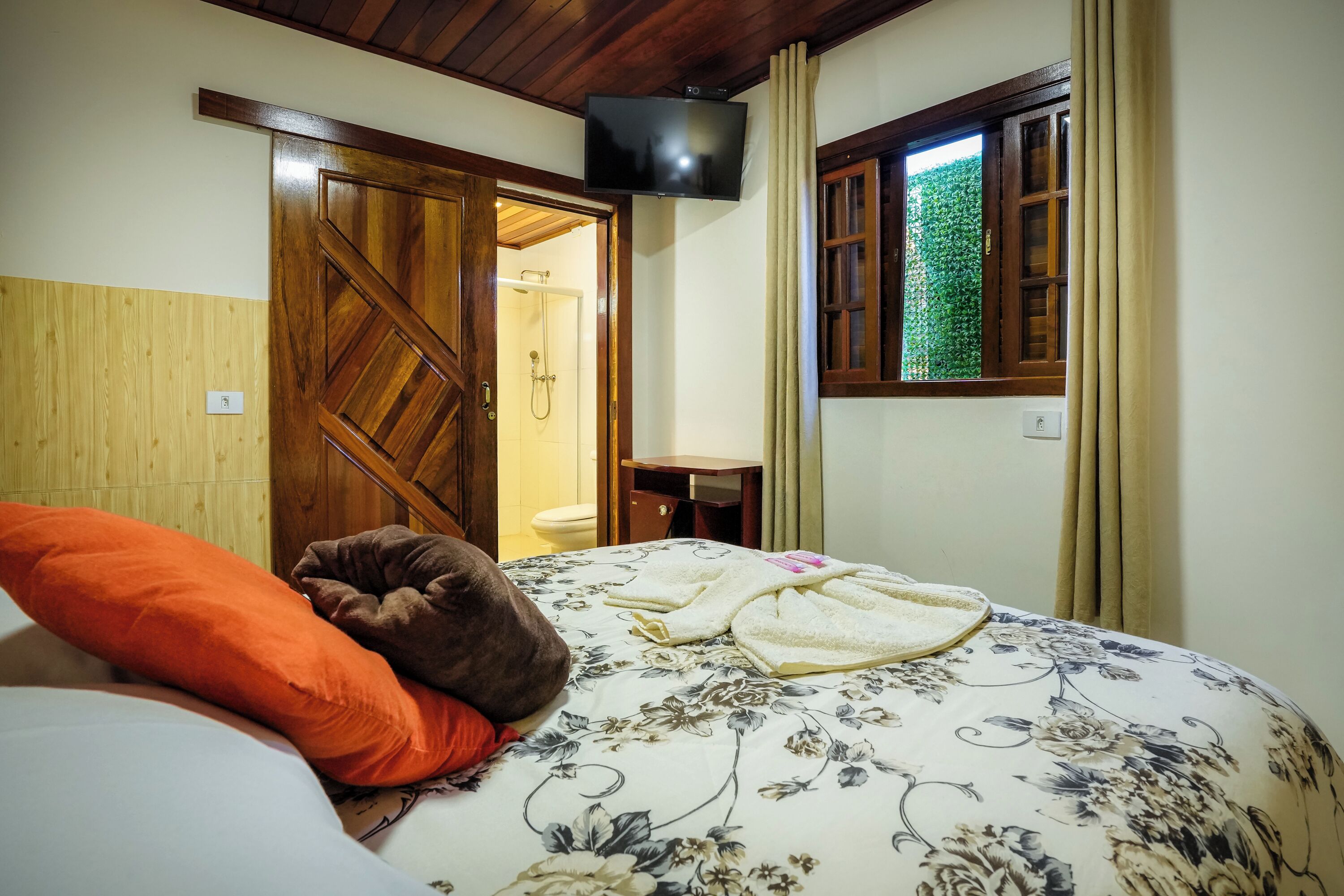 A bedroom with a wooden door, a shower, a small table, and a TV mounted on the wall.