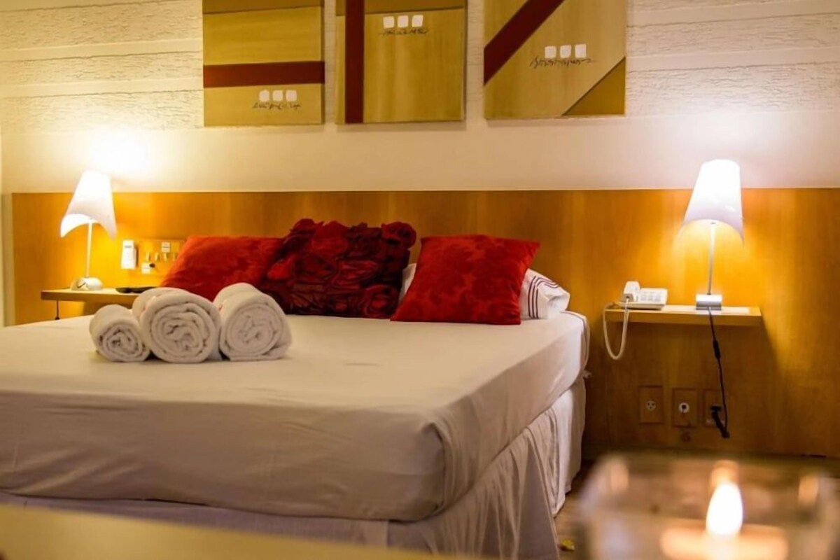 A hotel room with a neatly made bed, red pillows, and two bedside lamps.