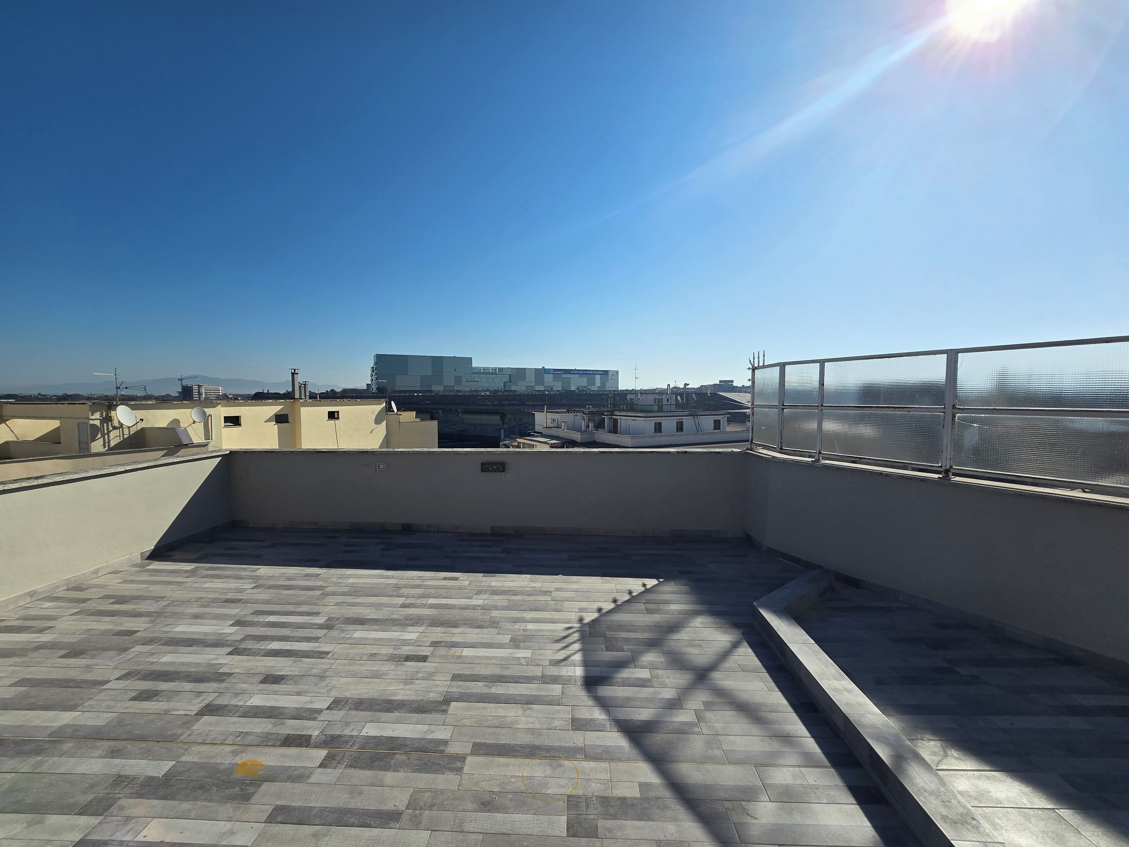 Une terrasse sur le toit, dotée d’un sol carrelé, d’une rambarde en métal et offrant une vue sur les bâtiments lointains.