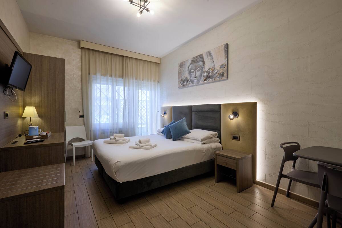 A hotel room with a bed, bedside tables, a desk, a chair, a TV, and a window with curtains.