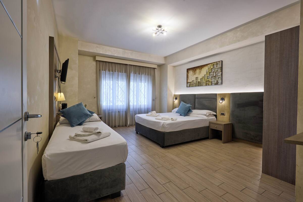 A hotel room with two beds, a large window with curtains, a painting on the wall, and a ceiling light.