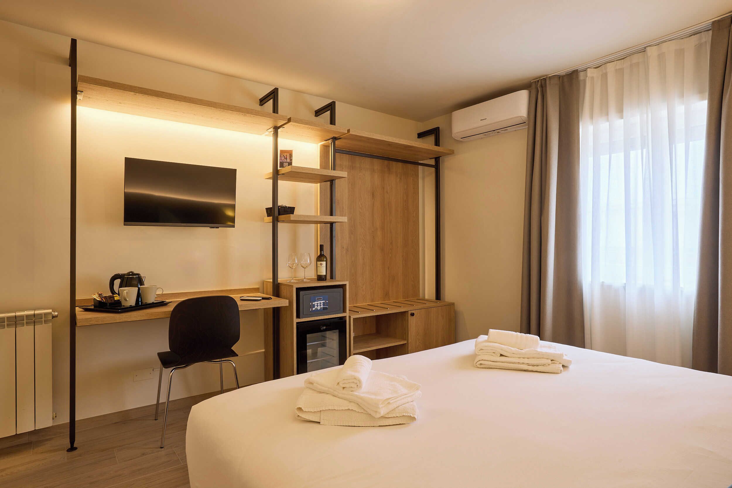 Een hotelkamer met een bed, een bureau met een computer, een stoel, een aan de muur bevestigde televisie en een raam met gordijnen.