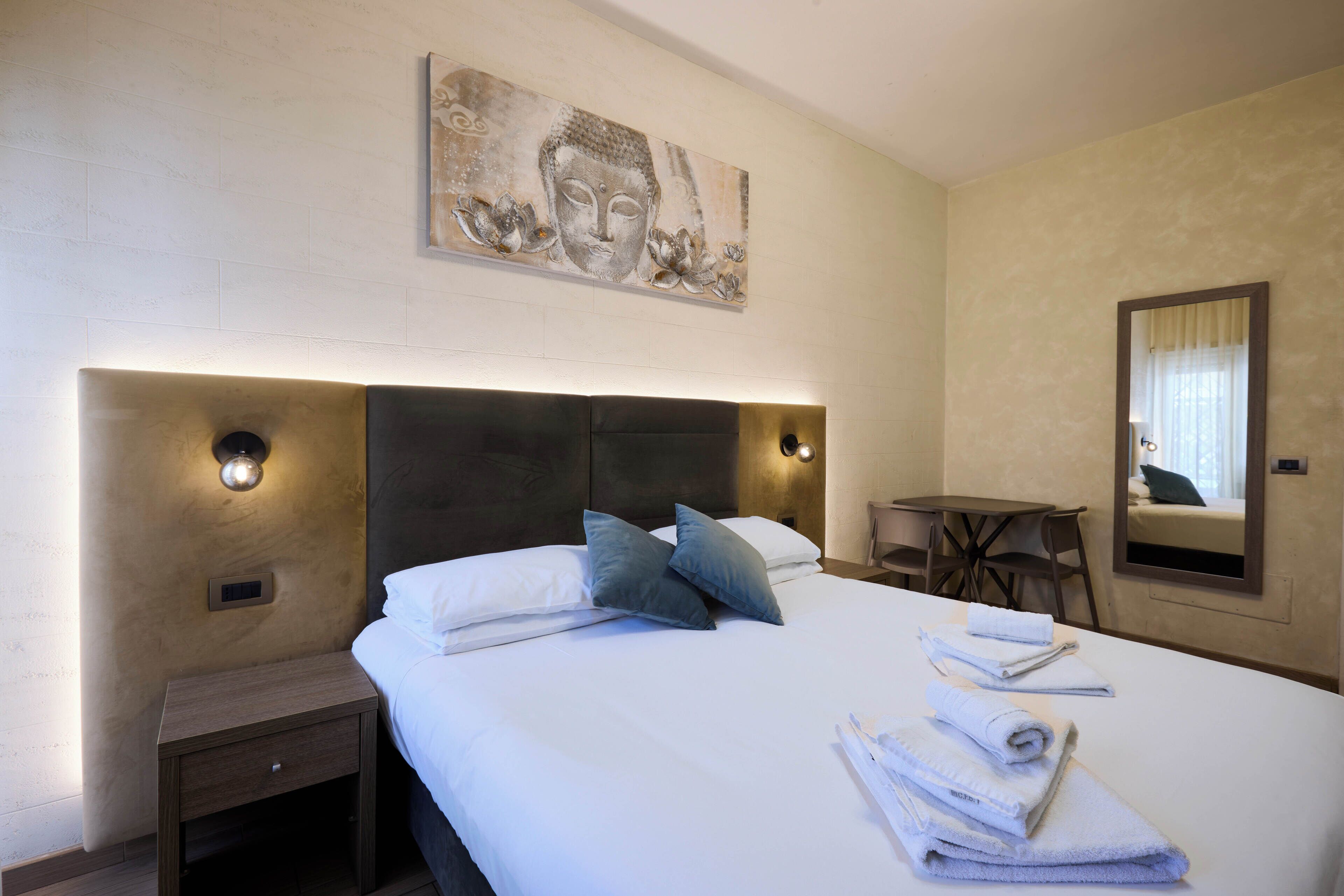 A hotel room with a large bed, two pillows, a headboard with integrated lighting, a small table, a chair, a mirror, and a wall-mounted artwork.