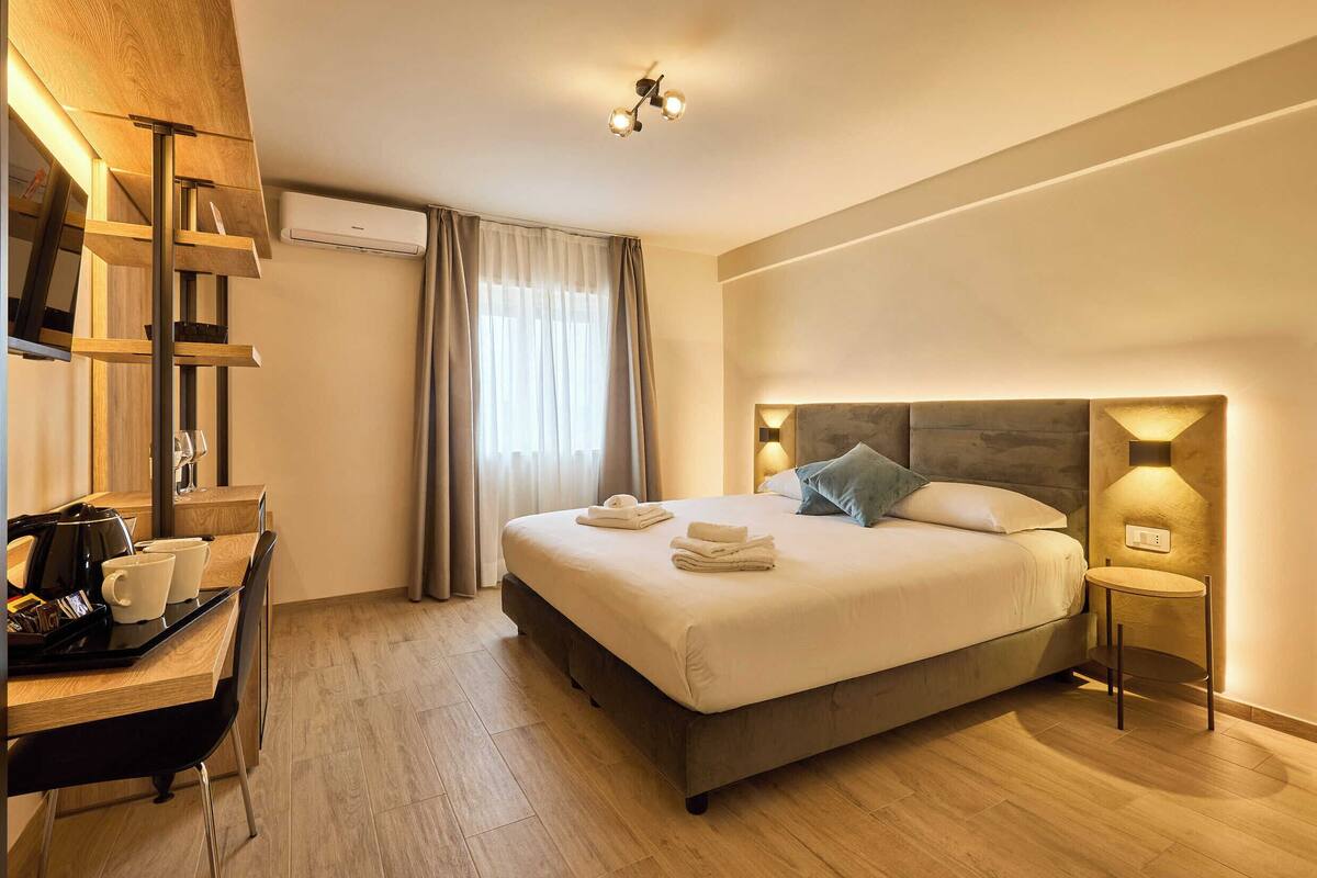 A modern hotel room with a large bed, bedside tables, a desk with a chair, and a window with curtains.