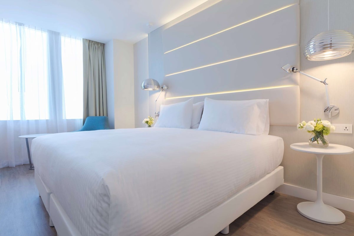 Una habitación de hotel moderna con una cama grande, lámparas de noche, una mesita con flores y vistas al exterior a través de la ventana.