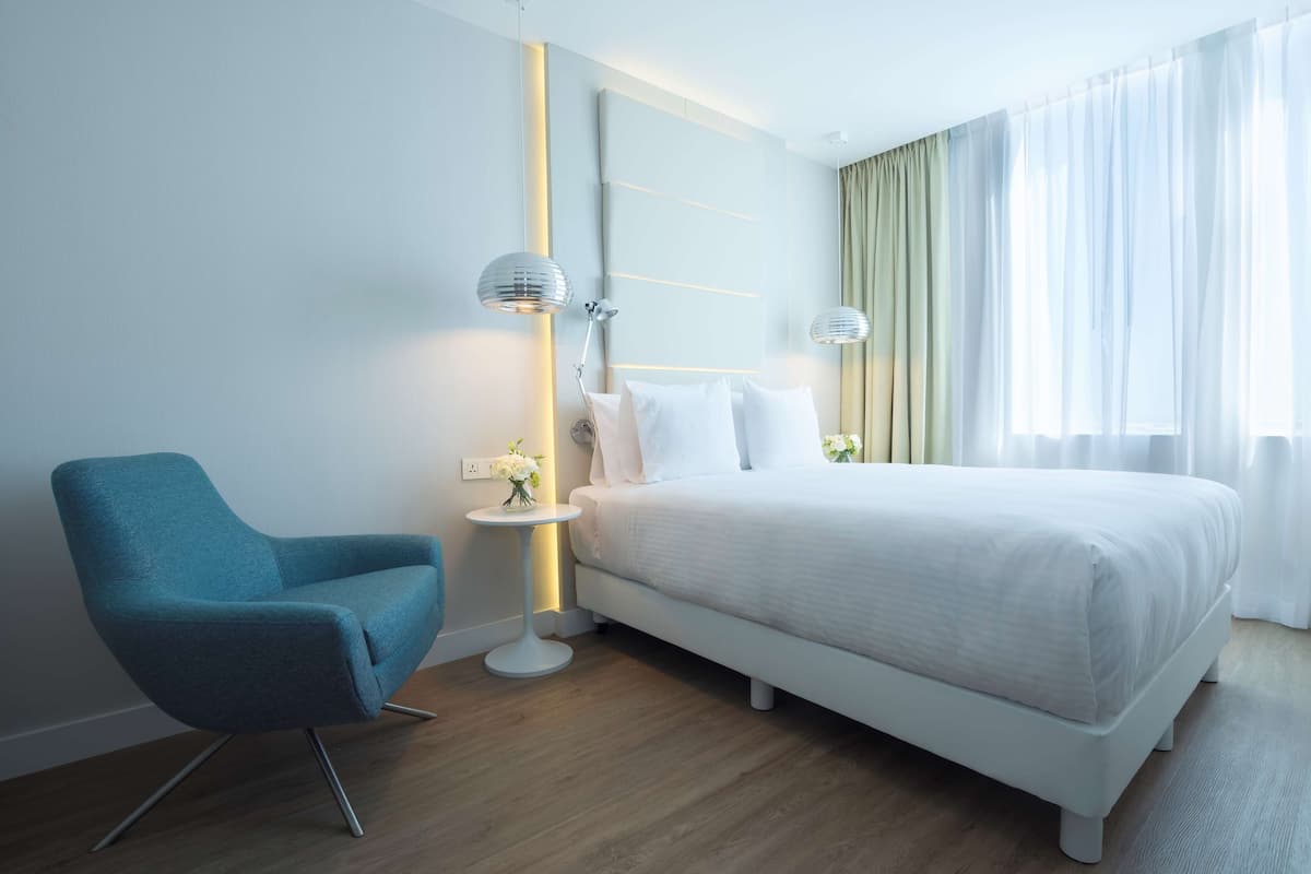 Una habitación de hotel moderna con una cama, un sillón azul, una mesita de noche con lámpara y un amplio ventanal con cortinas translúcidas.