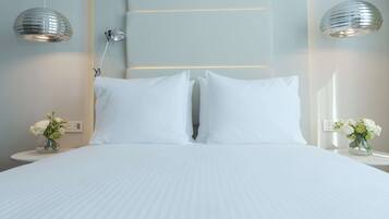 Una cama bien tendida con sábanas blancas, dos lámparas fijadas a la pared y dos mesitas de noche con arreglos florales.