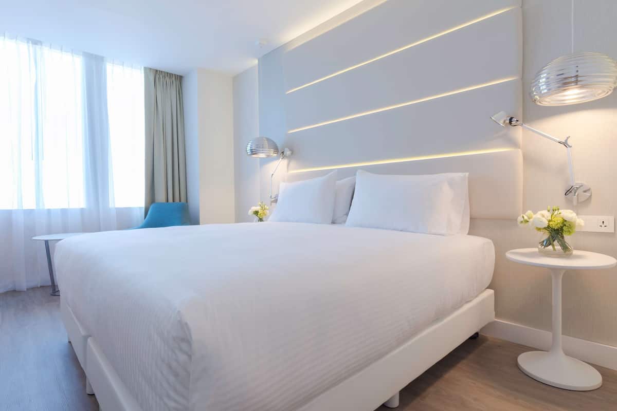 Habitación de hotel moderna con una cama grande, lámparas de noche, una mesita con un florero y vistas al exterior.