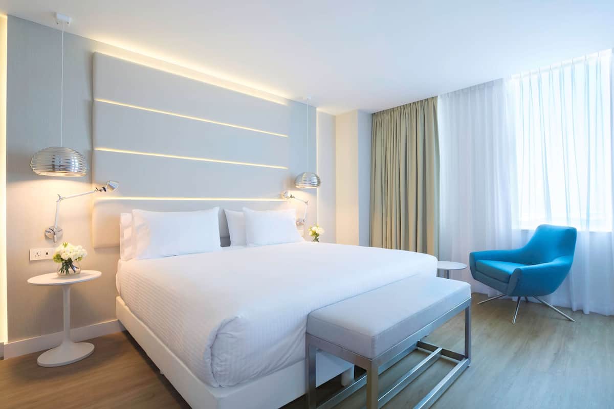 Habitación de hotel moderna con una cama grande, lámparas de noche, una mesita redonda, una silla azul y vistas al exterior.