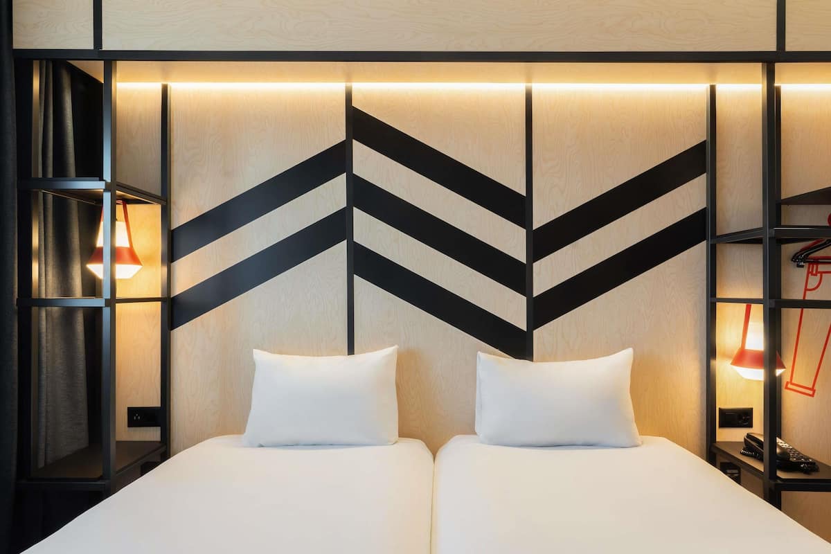 A hotel room with two beds, a modern geometric wall design, and a nightstand with a lamp.