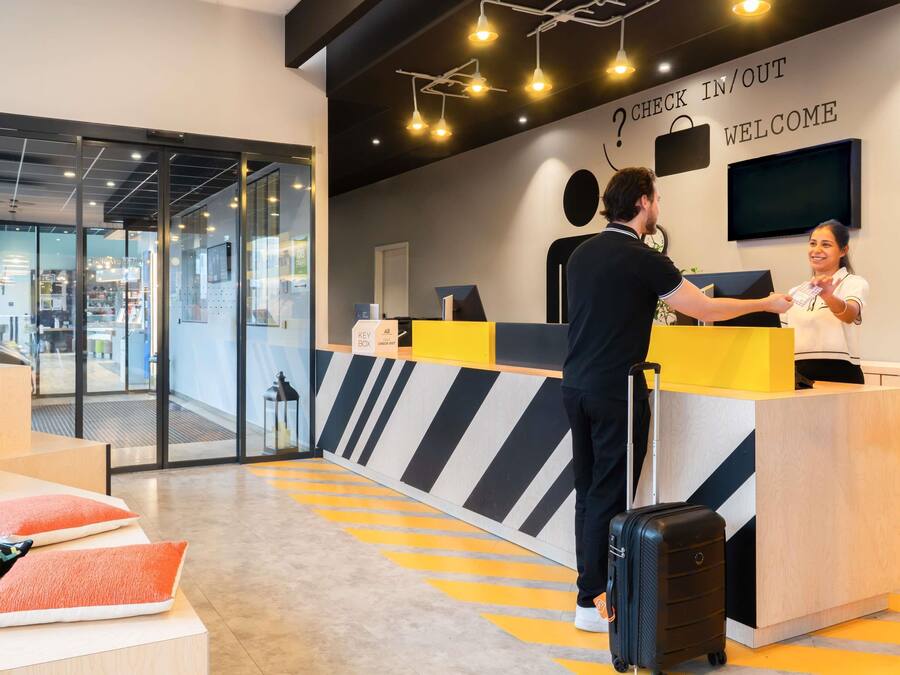 Un mostrador de recepción de hotel con un cartel de registro de entrada/salida, un televisor y un carrito de equipaje.
