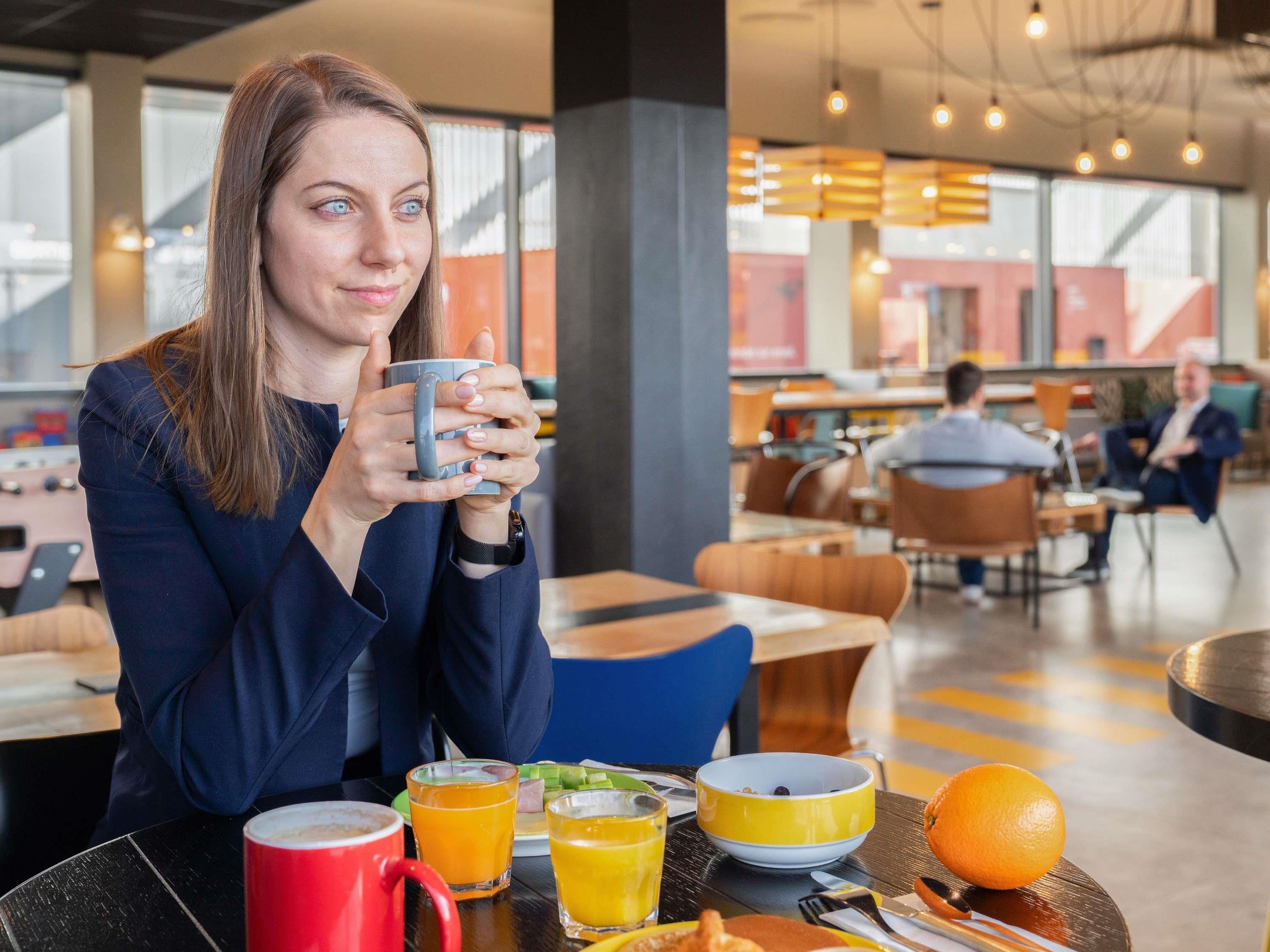 Uma pessoa sentada a uma mesa com uma chávena, uma tigela e um prato com comida, numa zona de refeições moderna com outros clientes ao fundo.
