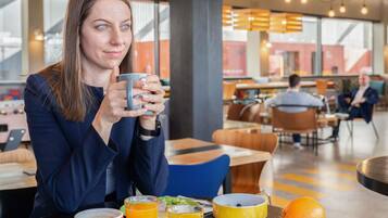 Uma pessoa sentada a uma mesa com uma chávena, uma tigela e um prato com comida, numa zona de refeições moderna com outros clientes ao fundo.