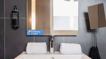 A bathroom with a white sink, two white towels, a mirror, a soap dispenser, and a sign indicating a drinking water tap.