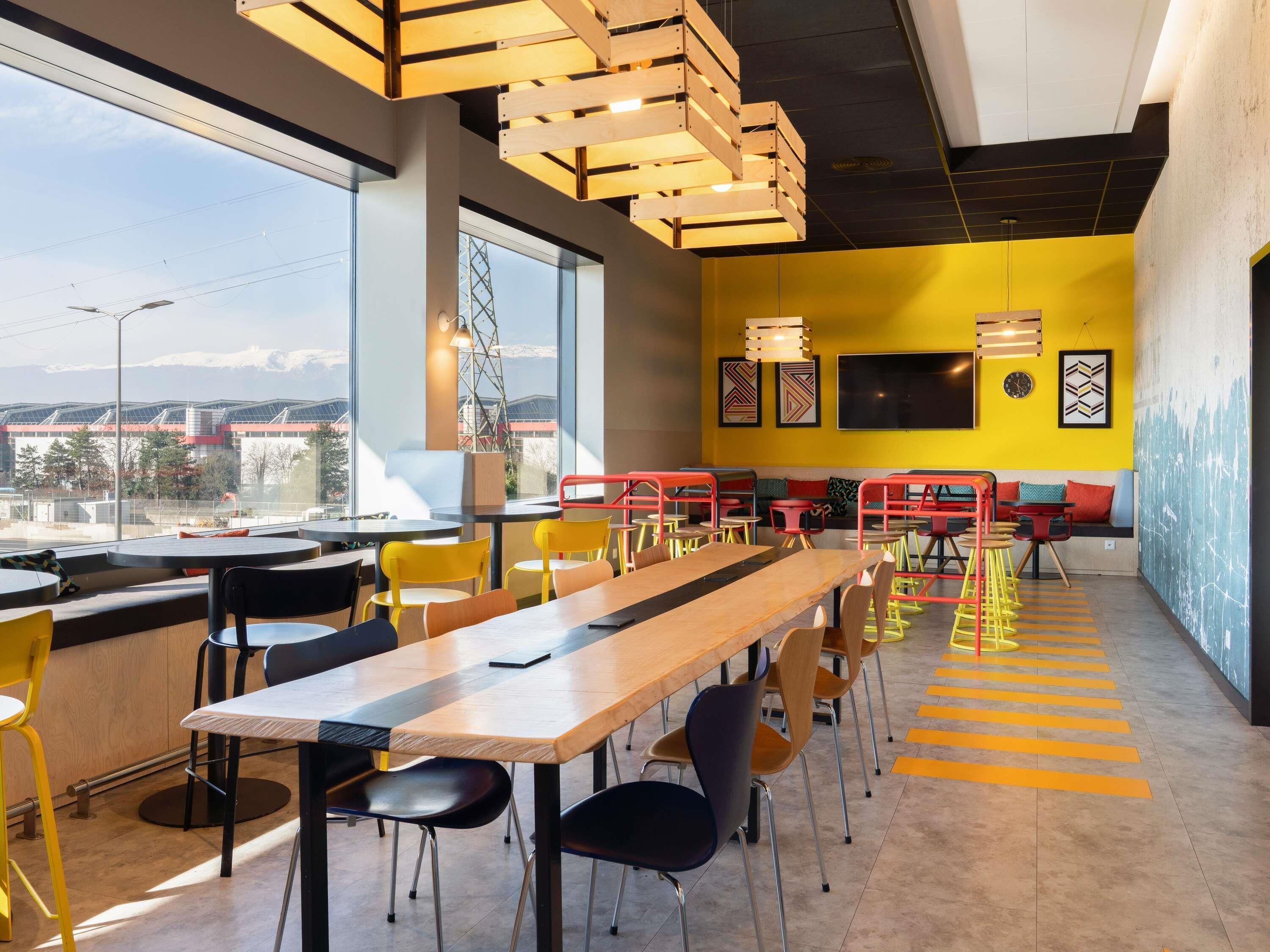 A modern dining area with a long wooden table, multiple chairs, and a yellow accent wall.