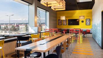 A modern dining area with a long wooden table, multiple chairs, and a yellow accent wall.