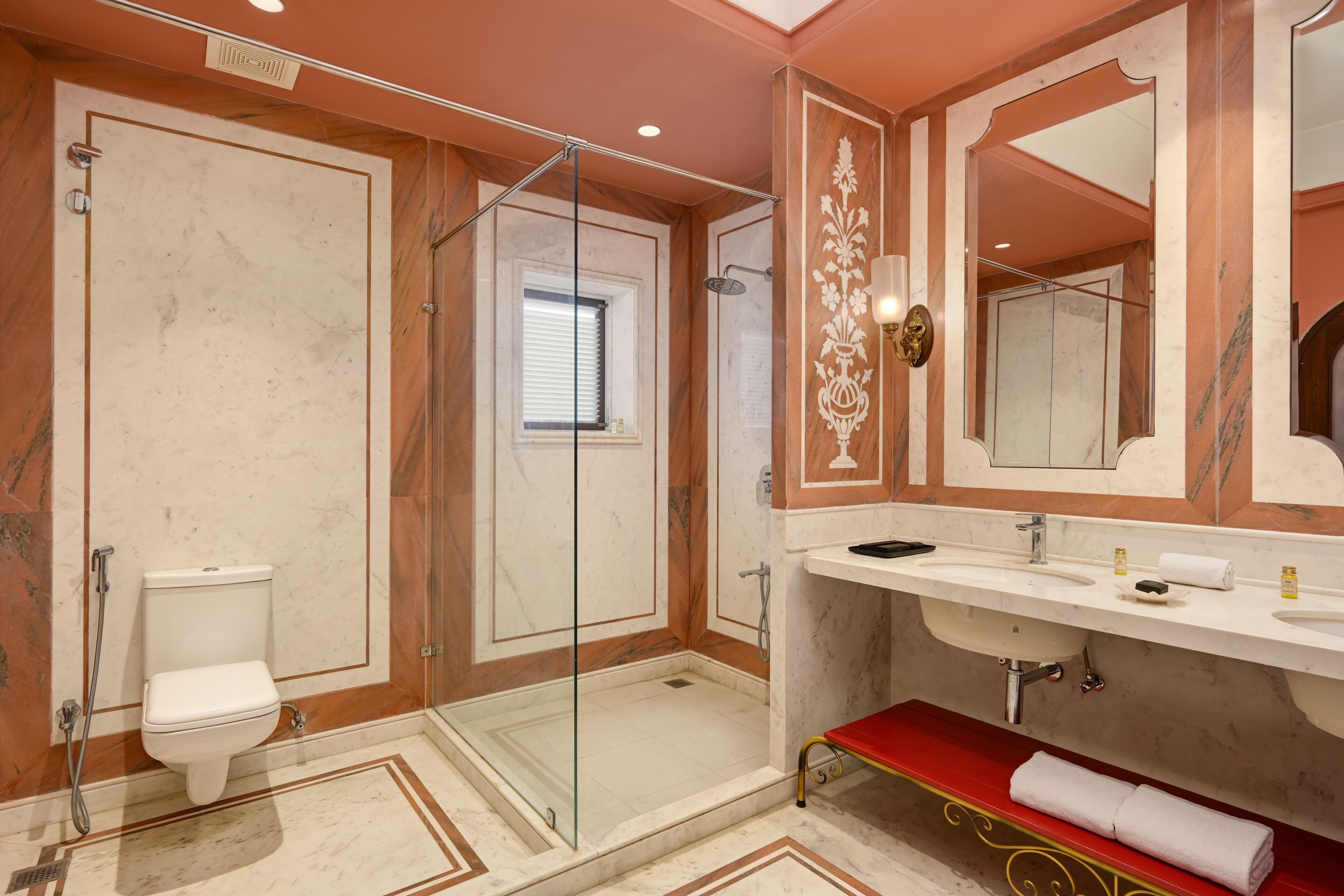 A bathroom with a glass-enclosed shower, a white toilet, a marble floor, and a red bench.