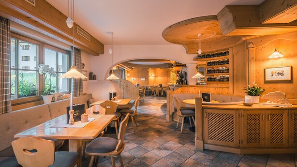A restaurant with wooden furniture, a bar area, and a view of the outside through large windows. - Garni Raetia (Corvara in Badia)