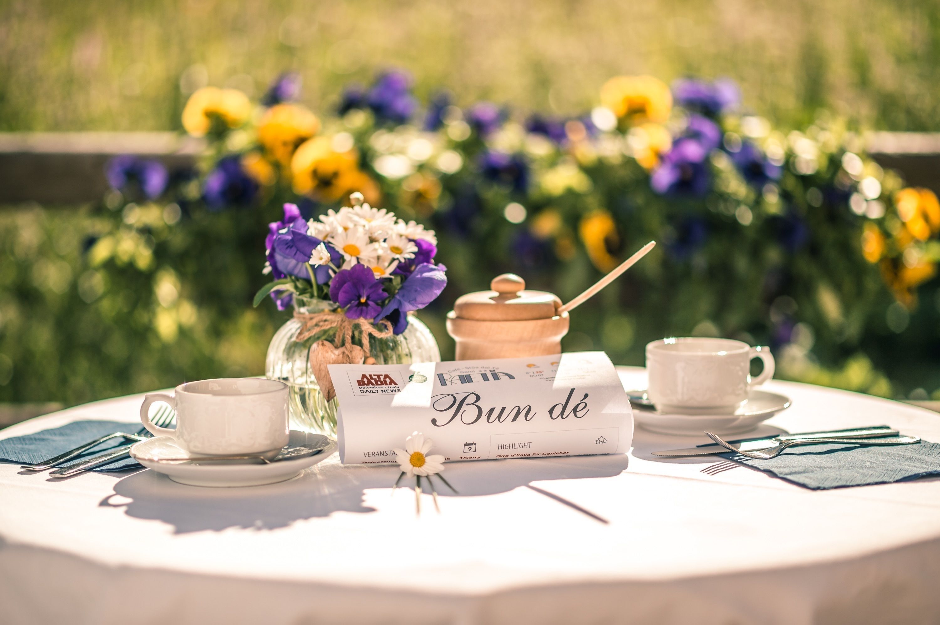 A table set outdoors with a book, two cups, a jar, and a flower.