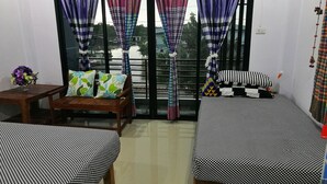 Una habitación con dos camas, una silla de madera, una mesita con un florero y un balcón con cortinas coloridas.
