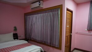 Un dormitorio con cama, ventana con cortinas cuadriculadas, equipo de aire acondicionado, puerta de madera y una planta pequeña.