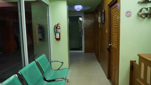 Un pasillo con sillas verdes, un extintor y una puerta con un cartel de prohibido fumar.