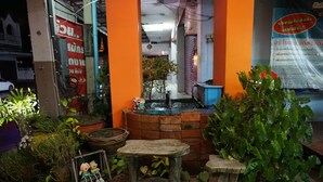 Un pequeño espacio al aire libre con un banco de madera, una mesa pequeña y una fuente de agua. Hay plantas en macetas y un letrero con texto en un idioma extranjero.