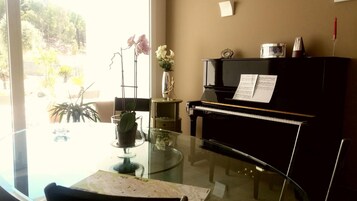 A room with a glass table, a piano, a vase with flowers, and a view of greenery outside.