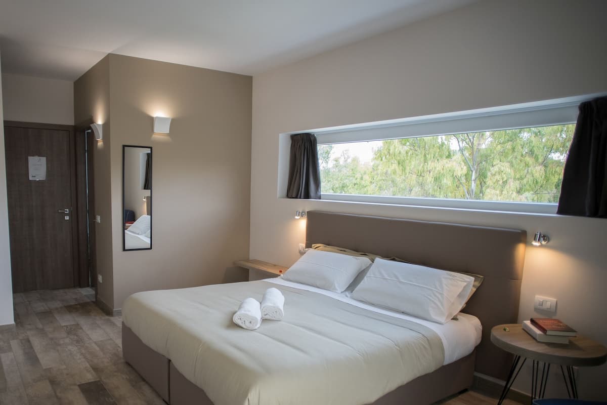Une chambre d’hôtel avec un lit, une table de chevet sur laquelle se trouve un livre, une fenêtre avec des rideaux et un miroir au mur.