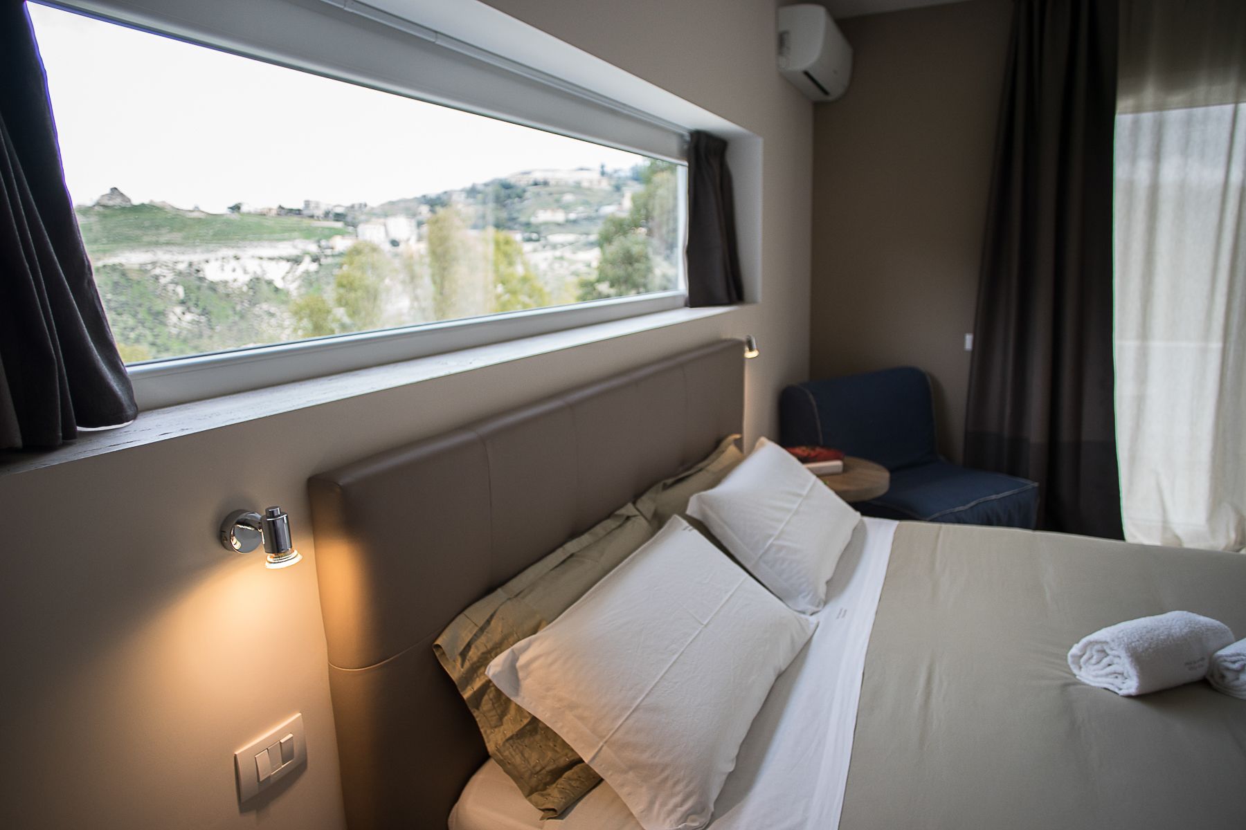 Une chambre d’hôtel comprenant un lit, une chaise, une fenêtre donnant sur la ville et un climatiseur fixé au mur.