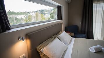 A hotel room with a bed, a chair, a window with a view of a cityscape, and a wall-mounted air conditioning unit.