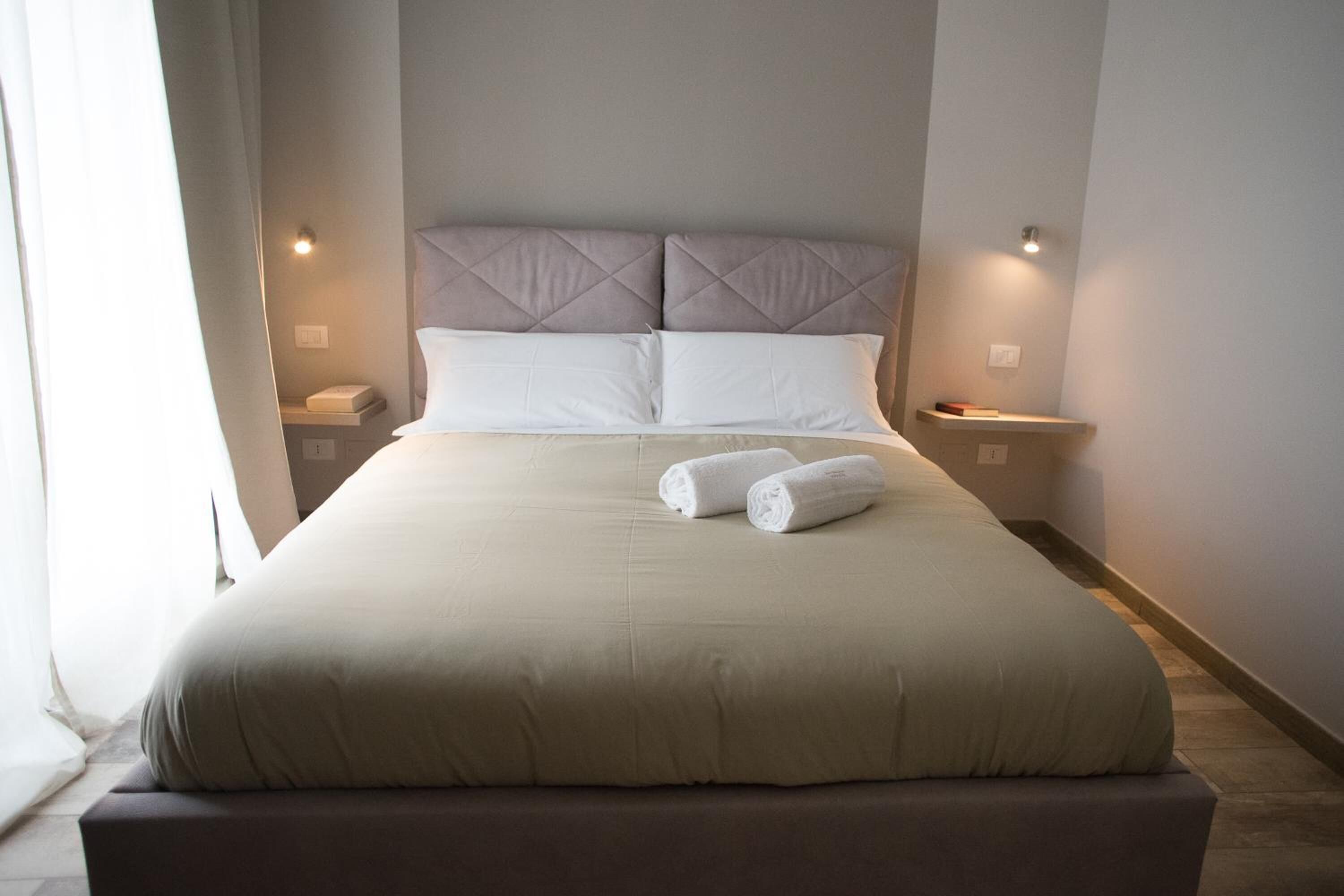 A neatly made bed with a headboard, two bedside tables, and a pair of rolled towels on the bed.